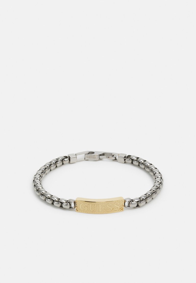 Pulseira de corrente de prata com uma placa retangular dourada gravada com "GUESS", segurada por um fecho, exibida sobre um fundo branco.