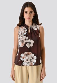 Donna che indossa un top senza maniche marrone scuro con stampa floreale e scollatura a chiave insieme a pantaloni beige, in piedi contro uno sfondo grigio chiaro uniforme.