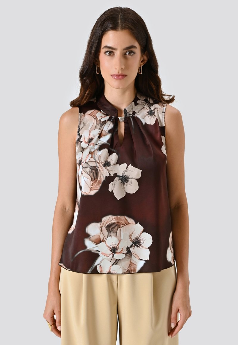 Donna che indossa un top senza maniche marrone scuro con stampa floreale e scollatura a chiave insieme a pantaloni beige, in piedi contro uno sfondo grigio chiaro uniforme.