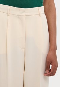 Kremowe, dopasowane spodnie wykonane z lekkiego materiału, z płaskim przodem, bocznymi kieszeniami i gładką teksturą, o średnim stanie.