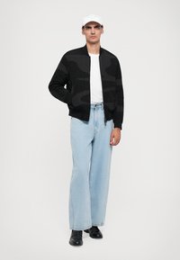 Black bomber jacket with textured patterns, white t-shirt, light blue wide-leg jeans, and black shoes; model wears a white cap.