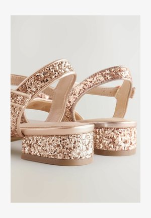 Rózsaarany szandálok platform talppal, csillogó felsőrésszel, széles pántokkal és sima, fényes sarokkal. A talpuk strapabíró és lapos.