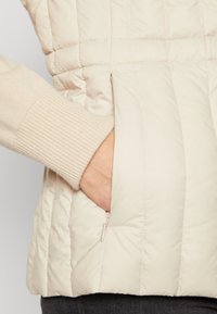 Pikowana, jasnobeżowa kamizelka z beżowym ściągaczem przy rękawach, wyposażona w boczną kieszeń na suwak. Gładka faktura i miękkie wykończenie.