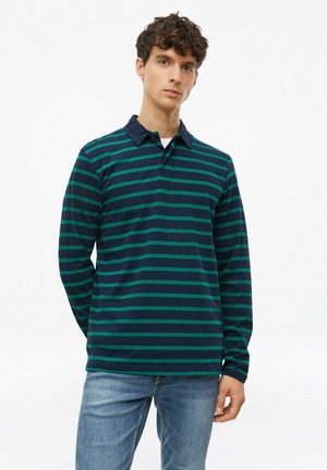 Jonge man die een donkerblauw en groen gestreepte polo met lange mouwen en lichtblauwe spijkerbroek draagt tegen een effen witte achtergrond.