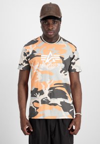 Man som bär en brun keps, pärlhalsband och armband, orange och grå kamouflage t-shirt från Alpha Industries, och svarta byxor mot vit bakgrund.