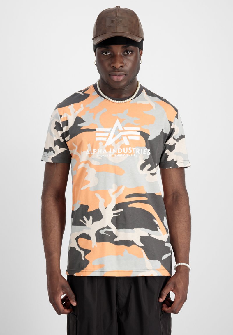 Man som bär en brun keps, pärlhalsband och armband, orange och grå kamouflage t-shirt från Alpha Industries, och svarta byxor mot vit bakgrund.