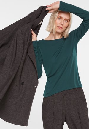 Frau mit blondem Haar trägt ein türkisfarbenes Langarmshirt und dunkelgraue Hose und hält einen passenden dunkelgrauen Blazer über der Schulter.