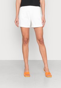 Femme portant un short blanc à ourlet roulé, un haut noir et des sandales à enfiler ouvertes aux orteils de couleur orange vif, se tenant devant un fond clair.
