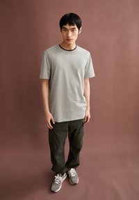 Jonge man in een lichtgrijs t-shirt, donkergekleurde cargo-broeken en grijze sneakers staat tegen een effen bruine achtergrond.