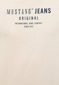 Beige cotton t-shirt with dark blue printed text reading "MUSTANG® JEANS ORIGINAL INTERNATIONAL JEANS COMPANY SINCE 1932." Smooth texture.