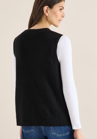 Schwarze Strickweste mit texturierter Oberfläche, ärmellosem Design und rundem Ausschnitt, getragen über einem weißen Langarmshirt.