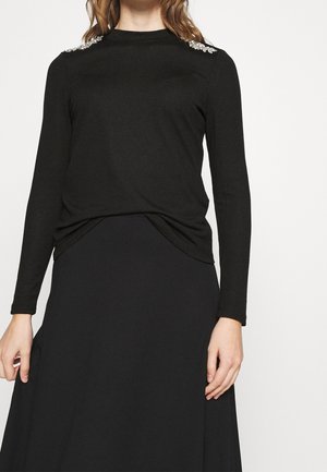 Kobieta ubrana w czarną bluzkę z długim rękawem ozdobioną srebrnymi koralikami na ramionach oraz czarną spódnicę.