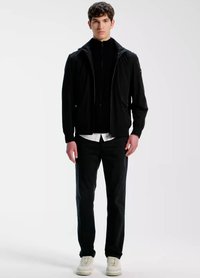 Črna jakna z zadrgo in kapuco, oblečena preko bele majice, kombinirana s črnimi hlačami in svetlo obarvanimi supergami. Gladek material, sodoben kroj.