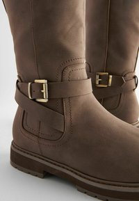 Bottes marron montant jusqu'au milieu du mollet avec une sangle décorative et une boucle en laiton, coutures visibles et semelle épaisse à crampons sur fond blanc.