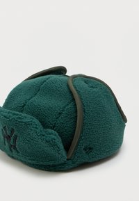 Zelená fleecová čapica so zaobleným tvarom, s vyšitým logom v čiernej farbe, kontrastným lemom a mäkkým štruktúrovaným povrchom.