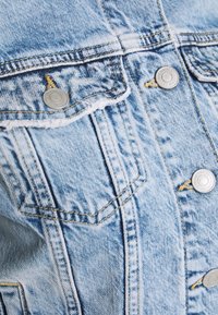 Vaaleansininen farkkutakki, jossa on nappikiinnitykset, kaksi rintataskua ja kontrastiväriset ompeleet. Kankaan pinta on hieman haalistunut.