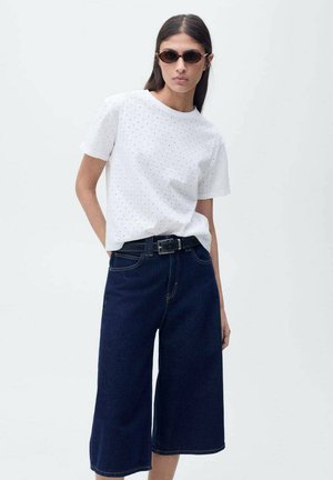 Chemise blanche à manches courtes avec de petits trous et culottes en denim larges de couleur bleu foncé, complétées par une ceinture noire et des lunettes de soleil ovales.