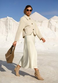 Woman wearing beige jacket, white skirt, and tan ankle boots, holding brown handbag, walking on white textured ground with white rock formations.