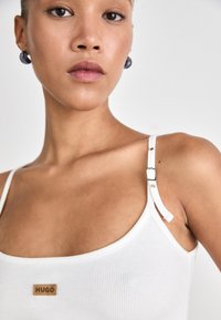 Valge soonikuline tank top reguleeritavate rihmadega. Omab väikest pruuni "HUGO" sildi ning sileda tekstuuri. Mudel kannab aksessuaare, nagu kõrvarõngad.