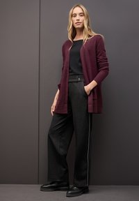 Cardigan largo burdeos con puños acanalados sobre una camisa negra, combinado con pantalones anchos negros con una franja lateral y botas negras.