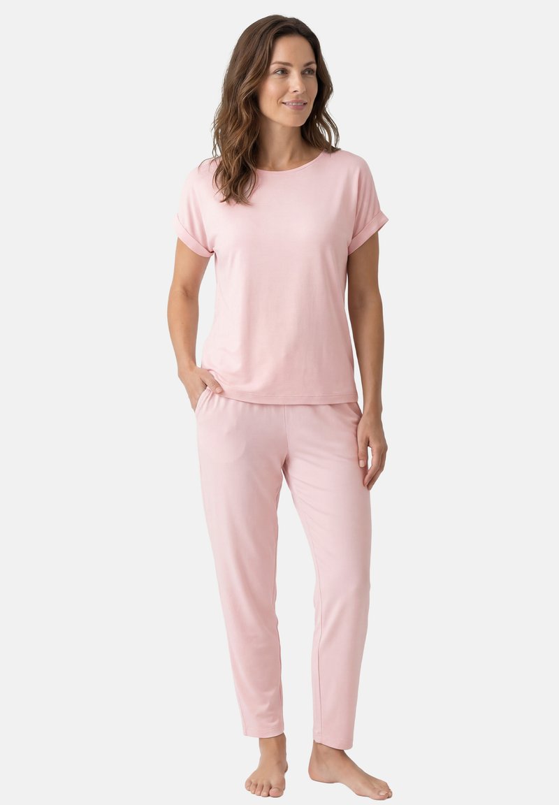 Femme debout pieds nus portant un haut à manches courtes rose clair et un pantalon assorti, les mains dans les poches, regardant légèrement sur le côté avec un sourire.