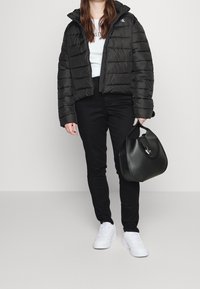 Černá péřová bunda se zipem a kapucí, v kombinaci s bílou košilí a černými džíny. Černá kabelka s elegantním designem a stříbrným akcentem. Bílé tenisky.
