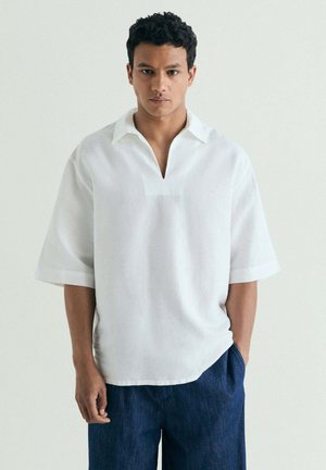 Hombre de pie mirando al frente, lleva una camisa blanca holgada con escote abierto y pantalones azul oscuro sobre un fondo claro y liso.