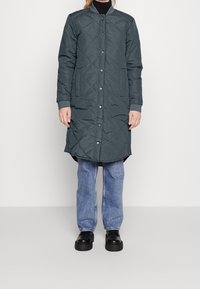 Fodrad långjacka i mörkgrön med ståkrage, tryckknappar och sidofickor, tillsammans med ljusblå jeans och svarta skor.