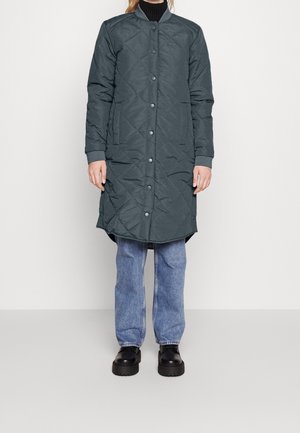 Vatteret lang jakke i mørkegrøn med ståkrave, trykknapper og sidelommer, parret med lyseblå jeans og sorte sko.