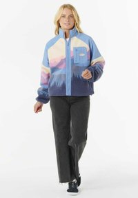 Kvinde går iført en lynlås fleecejakke med et landskabsdesign i blå, lilla og creme, sorte brede bukser og sorte sneakers.