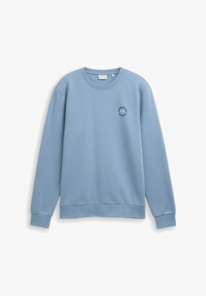 Sweatshirt léger bleu clair à manches longues et encolure crewneck, avec des poignets et un ourlet côtelés, et un petit logo circulaire sur la poitrine gauche indiquant "Tom Tailor Team 1962."