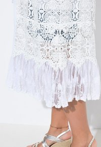Abito bianco in pizzo con intricate fantasie floreali, dotato di un orlo a strati con volant. Indossato con sandali argentati con cinturini e un braccialetto alla caviglia multicolore.