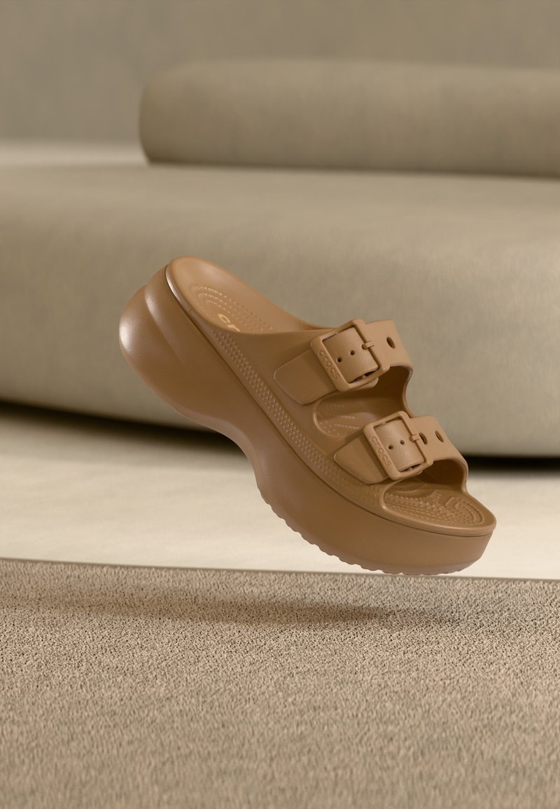 Καφέ πλατφόρμα σανδάλι με δύο ρυθμιζόμενους ιμάντες με αγκράφα, υφασμένο πάτο και χοντρή σόλα που αιωρείται πάνω από μπεζ χαλί.