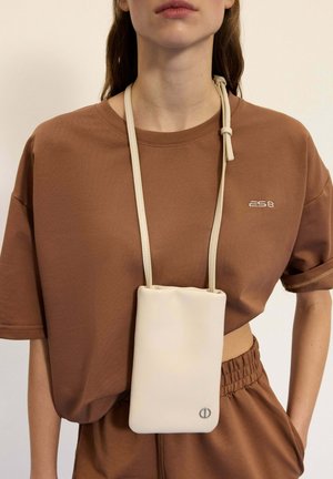 Beżowa torba na smartfona wykonana z gładkiego materiału, z cienkim paskiem. Modelka ma na sobie brązowy oversize'owy T-shirt z delikatnym detalem logo.