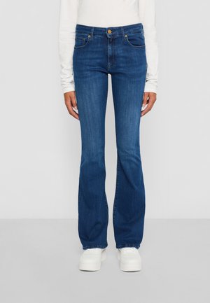 Person med blå bootcut jeans, hvit langermet topp og hvite sneakers, som står mot en ensfarget lys grå bakgrunn.