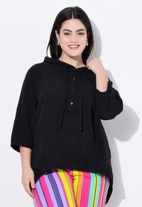 Felpa nera oversize con cappuccio, in tessuto texturizzato, con tre bottoni sulla parte frontale e un orlo curvo, abbinata a pantaloni a righe multicolore.