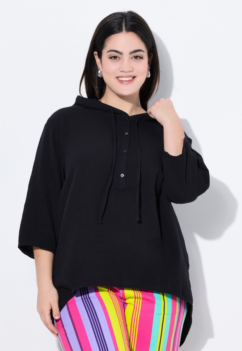 Felpa nera oversize con cappuccio, in tessuto texturizzato, con tre bottoni sulla parte frontale e un orlo curvo, abbinata a pantaloni a righe multicolore.
