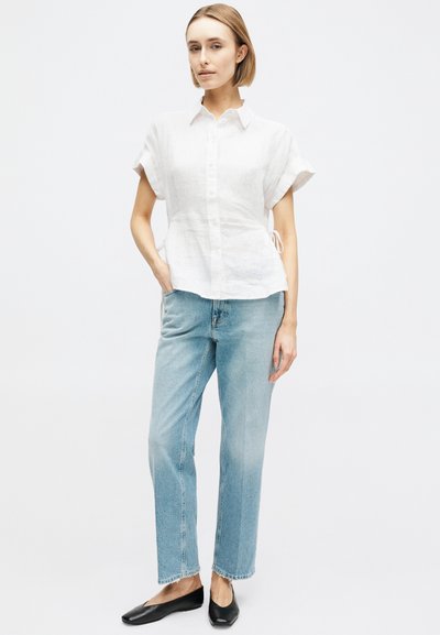 Γυναίκα που στέκεται με το ένα χέρι στην τσέπη, φοράει λευκό πουκάμισο με κοντά μανίκια και κουμπιά, ανοιχτόχρωμο μπλε τζιν και μαύρα μπαλαρίνες.