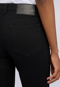 Černé džínové kalhoty s zadní kapsou, jemným švem a obdélníkovou koženou nášivkou s nápisem "TRUSSARDI." Hladká textura.