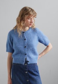 Vaaleansininen lyhythihainen neulecardigan, jossa on pehmeä pinta, mustat napit ja kaksi etutaskua, yhdistettynä raidalliseen farkkuhameeseen.