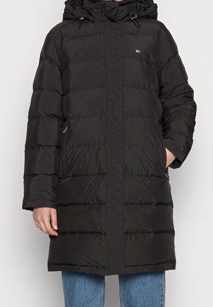 Manteau d'hiver long noir matelassé avec capuche, poches zippées et un petit logo sur la poitrine, porté avec un jean bleu.
