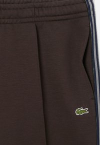 Pantaloni della tuta marroni in materiale morbido, con vita regolabile con coulisse, tasche laterali e una toppa con logo verde raffigurante un coccodrillo.