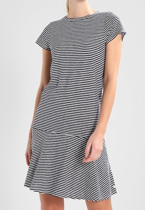 Kvinde iført en sort og hvid kortærmet kjole med vandrette striber og en udvidet nederdel, stående mod en ensartet lys baggrund.