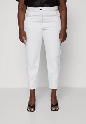 Personne portant un pantalon blanc taille haute à jambes droites aux chevilles et des sandales noires à talons ouvertes aux orteils, debout devant un fond uni.