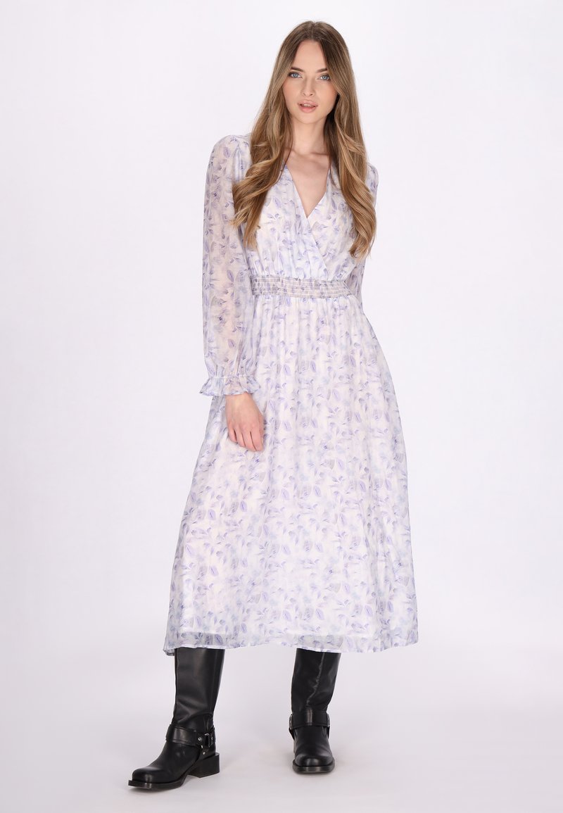 Helles lila Blumenkleid mit langen Ärmeln, das Rüschenmanschetten und eine geraffte Taille aufweist, kombiniert mit knielangen schwarzen Stiefeln.