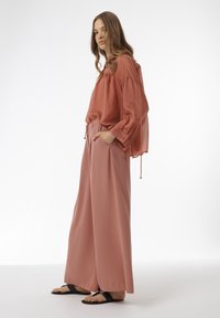 Roestkleurige blouse met trekkoorddetails, gecombineerd met losse, wijde broekspijpen. Een zachte stoffen textuur en zwarte platte sandalen complementeren de look.