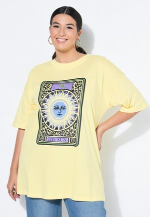 Hymyilevä tummahiuksinen nainen pukeutuu ylisuurikokoiseen keltaiseen T-paitaan, jossa on "Aurinko"-tarottikortin kuva ja siniset farkut.