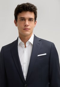 Giacca di abito blu navy con rever a notched, chiusura a un bottone e camicia bianca sotto. Il fazzoletto da taschino bianco valorizza la giacca.