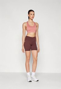 Reggiseno sportivo rosa chiaro e pantaloncini aderenti bordeaux scuro. Il tessuto appare liscio con cuciture minime e senza motivi visibili. Scarpe da ginnastica bianche.