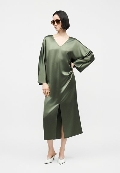 Γυναίκα με μακρύ πράσινο σατέν φόρεμα V-λαιμόκοψη με σκίσιμο μπροστά, άσπρα μυτερά τακούνια και καφέ γυαλιά ηλίου, ποζάρει με το χέρι στη μέση.